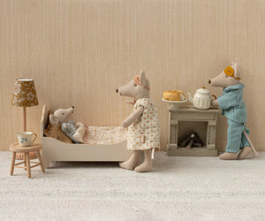 Three Maileg mice in a miniature bedroom: one snuggled in the Maileg Bed, Mouse - Off White, one standing nearby, and another in pajamas making tea and snacks on a tiny stove.
