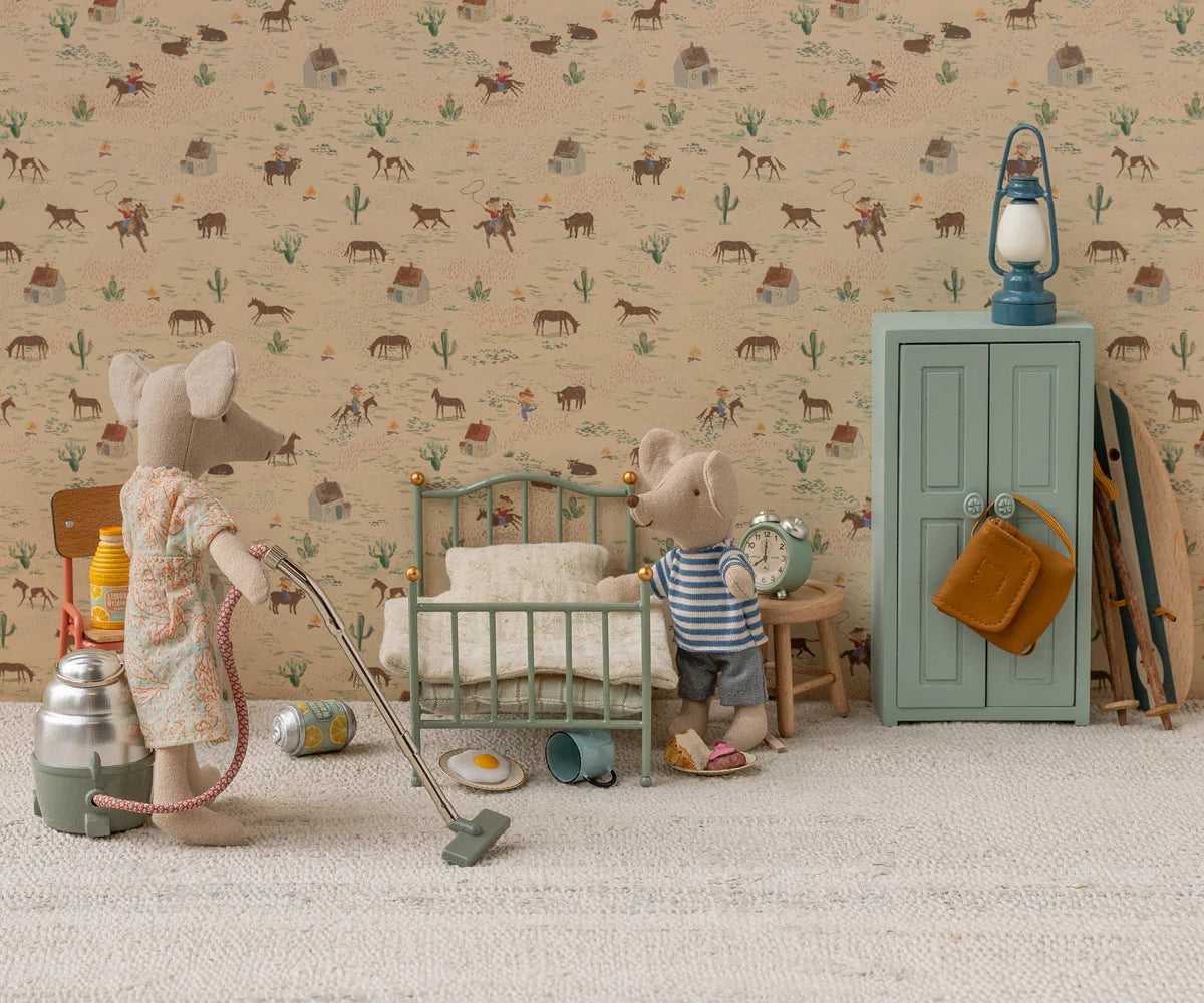 Two toy mice in a miniature room: one vacuums while the other stands by a Maileg Vintage Single Bed, Mouse - Mint. The scene features Maileg bedding, a green cabinet, scattered items, and wallpaper with western-themed illustrations.