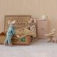 Maileg’s "Mum and Dad in Cigarbox" features two toy mice standing beside an open cigar box bed, complete with a soft mattress and blanket. Surrounding them are a pink cabinet, a round table with cake, a teapot, and a small stool on a light-colored carpeted floor.