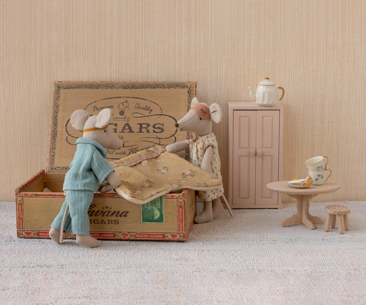 Maileg’s "Mum and Dad in Cigarbox" features two toy mice standing beside an open cigar box bed, complete with a soft mattress and blanket. Surrounding them are a pink cabinet, a round table with cake, a teapot, and a small stool on a light-colored carpeted floor.