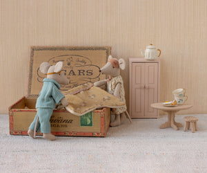 Maileg’s "Mum and Dad in Cigarbox" features two toy mice standing beside an open cigar box bed, complete with a soft mattress and blanket. Surrounding them are a pink cabinet, a round table with cake, a teapot, and a small stool on a light-colored carpeted floor.
