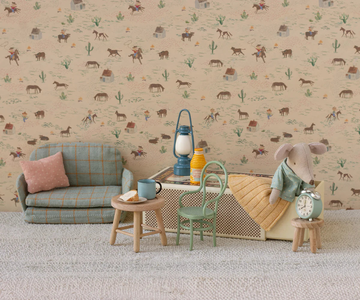 A charming miniature living room featuring Maileg’s Vintage Chair, Mouse - Mint, with tiny furniture, a toy mouse, lamp, clock, cups, bottle, and bread slice—all set against a patterned wall and carpeted floor.