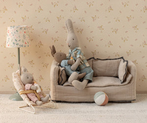 Three micro-sized Maileg rabbits relax in a dollhouse living room, featuring a sofa, lamp, soft toy ball, and the Maileg Babysitter, Micro - Off White.