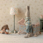 Two stuffed toy rabbits are in a nursery; one stands by the Maileg Cot Bed, Micro – Powder, while the other sits on the floor with a bowl. Nearby are a lamp, mug, bottle, and small fireplace—perfect for their micro-sized friends to gather.