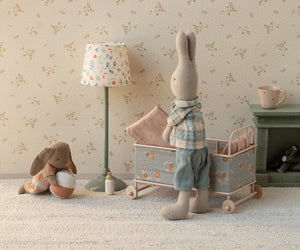 Two stuffed toy rabbits are in a nursery; one stands by the Maileg Cot Bed, Micro – Powder, while the other sits on the floor with a bowl. Nearby are a lamp, mug, bottle, and small fireplace—perfect for their micro-sized friends to gather.