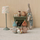 Three Maileg stuffed rabbits in knit clothes sit by a Maileg Fireplace, Miniature - Dusty Green, with milk bottles, watched over by a larger rabbit; a lamp and ball complete this cozy miniature home scene.