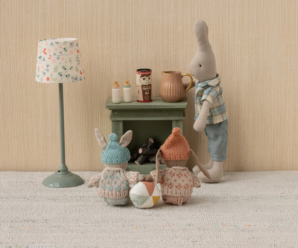 Three Maileg stuffed rabbits in knit clothes sit by a Maileg Fireplace, Miniature - Dusty Green, with milk bottles, watched over by a larger rabbit; a lamp and ball complete this cozy miniature home scene.