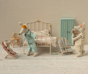 Three Maileg My-size mice in a miniature bedroom: one in bed, the Babysitter (My - Rose) holding a baby mouse, and another mouse in a bunny chair, with a clock, cabinet, and folded bedding in the background.