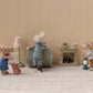 Three Maileg toy mice—one being the Hiker Mouse, Big Brother - Stripes—gather in a cozy room featuring a lamp, armchair, suitcase, fireplace, and tiny treats resting on the mantel. The adventurous Hiker Mouse has unrolled his sleeping bag by the fire, ready to rest after his imaginary journey.