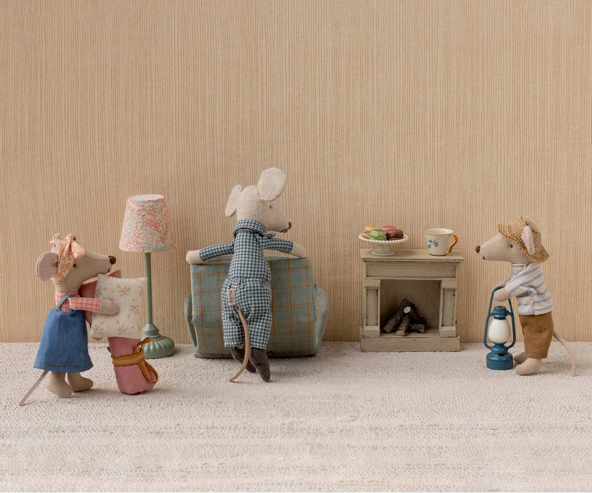 Three Maileg toy mice—one being the Hiker Mouse, Big Brother - Stripes—gather in a cozy room featuring a lamp, armchair, suitcase, fireplace, and tiny treats resting on the mantel. The adventurous Hiker Mouse has unrolled his sleeping bag by the fire, ready to rest after his imaginary journey.