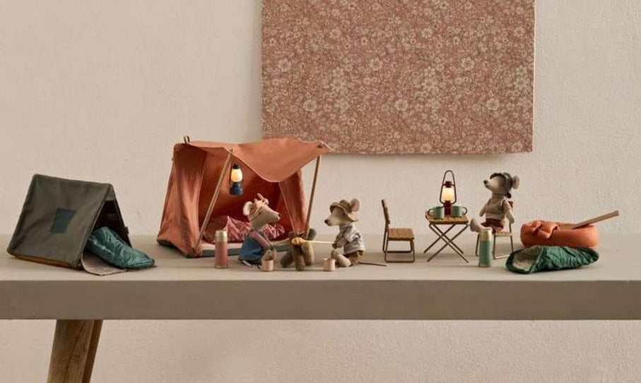 Four small Maileg mouse figurines have set up a delightful camping scene on a table, featuring tents—including the cheerful Happy Camper Tent - Mouse—along with sleeping bags, lanterns, and miniature camping accessories. This whimsical arrangement sparks imaginative play and perfectly captures the joy of outdoor adventures in charming, miniature detail.