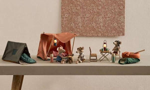 Four small Maileg mouse figurines have set up a delightful camping scene on a table, featuring tents—including the cheerful Happy Camper Tent - Mouse—along with sleeping bags, lanterns, and miniature camping accessories. This whimsical arrangement sparks imaginative play and perfectly captures the joy of outdoor adventures in charming, miniature detail.