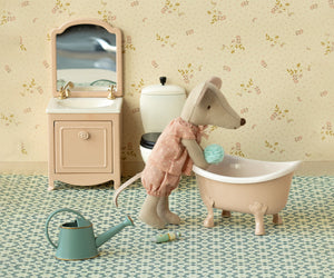 A Maileg Rose Bathtub – Mouse features a plush mouse in pajamas beside a vintage-inspired bathtub in the charming Mouse Hole Farmhouse bathroom, complete with a sink, mirror, toilet, watering can, and bottle on a patterned floor—perfect for the whole mouse family.