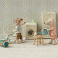 Two toy mice from Maileg are set in a cozy room with a washing machine, chair, rolled blanket, and stool, each showing off their delightful accessories. One mouse holds a watering can, while the other adds charm to the scene with its stylish hat and striped shirt. Their blue striped bag reveals a Toothbrush Set—perfect for whimsical bedtime routines—while patterned wallpaper brings this charming setting to life.