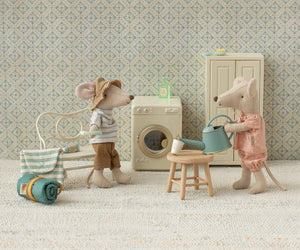 Two toy mice from Maileg are set in a cozy room with a washing machine, chair, rolled blanket, and stool, each showing off their delightful accessories. One mouse holds a watering can, while the other adds charm to the scene with its stylish hat and striped shirt. Their blue striped bag reveals a Toothbrush Set—perfect for whimsical bedtime routines—while patterned wallpaper brings this charming setting to life.