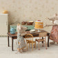 Two Maileg toy mice in dresses stand by a table topped with the Off White Party Cake and Cake Stand - Mouse, surrounded by miniature dishes and a teapot in a charming farmhouse kitchen furnished with tiny chairs, a fridge, and cabinets—perfect for delightful birthday celebrations.