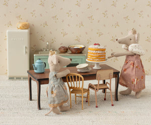 Two Maileg toy mice in dresses stand by a table topped with the Off White Party Cake and Cake Stand - Mouse, surrounded by miniature dishes and a teapot in a charming farmhouse kitchen furnished with tiny chairs, a fridge, and cabinets—perfect for delightful birthday celebrations.