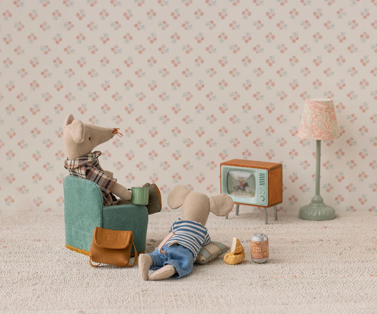 Two toy mice enjoy a cozy evening at Mouse Hole Farmhouse; one relaxes in a chair with a mug while the other lounges on the floor with snacks. Surrounded by charming miniature mouse accessories from Maileg, including their Television - Mouse, as well as a lamp and bag, they unwind together against a floral patterned backdrop.