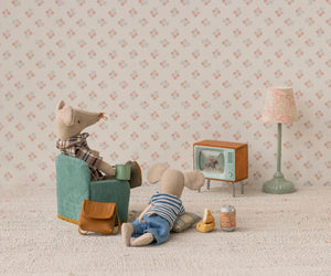 Two toy mice enjoy a cozy evening at Mouse Hole Farmhouse; one relaxes in a chair with a mug while the other lounges on the floor with snacks. Surrounded by charming miniature mouse accessories from Maileg, including their Television - Mouse, as well as a lamp and bag, they unwind together against a floral patterned backdrop.