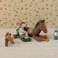 A Maileg stuffed mouse in camping gear sits on a sleeping bag beside a plush horse with a carrot, accompanied by the Blue Vintage Lantern - Mouse, and fishing gear, all displayed on a textured surface for charming farmhouse appeal.