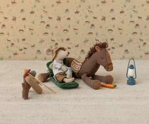 A Maileg stuffed mouse in camping gear sits on a sleeping bag beside a plush horse with a carrot, accompanied by the Blue Vintage Lantern - Mouse, and fishing gear, all displayed on a textured surface for charming farmhouse appeal.