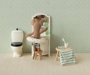 Beach Mouse with Yellow Dot Float - Little Brother by Maileg relaxes at the dollhouse vanity, dressed in playful swim trunks and his cheerful yellow dot float. With bunny ears perked up, he sits beside the toilet and a stool draped with a striped towel and cup—ready for any adventure, whether it’s bath time fun or imaginative beach play.