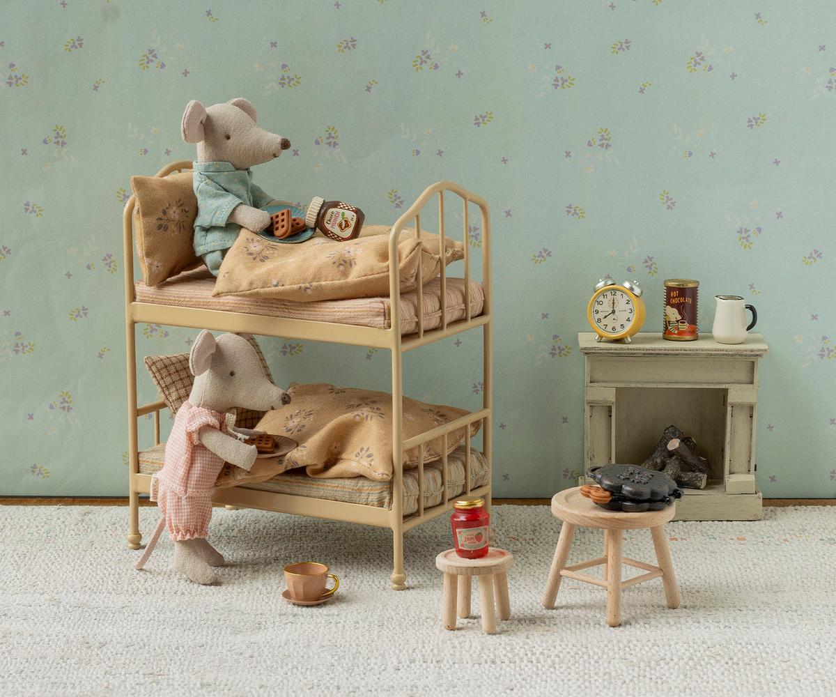 Two Maileg mice with books enjoy a cozy sleepover on the Wheat Bunk Bed - Mouse in a miniature Mouse Hole Farmhouse room, surrounded by a fireplace, clock, cups, and charming small furniture.