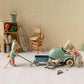 Three Maileg mice in a room with camping gear inspect a Light Mint Car – Mouse; one mouse lies under the car, one looks inside, and one pulls a pull along wagon.