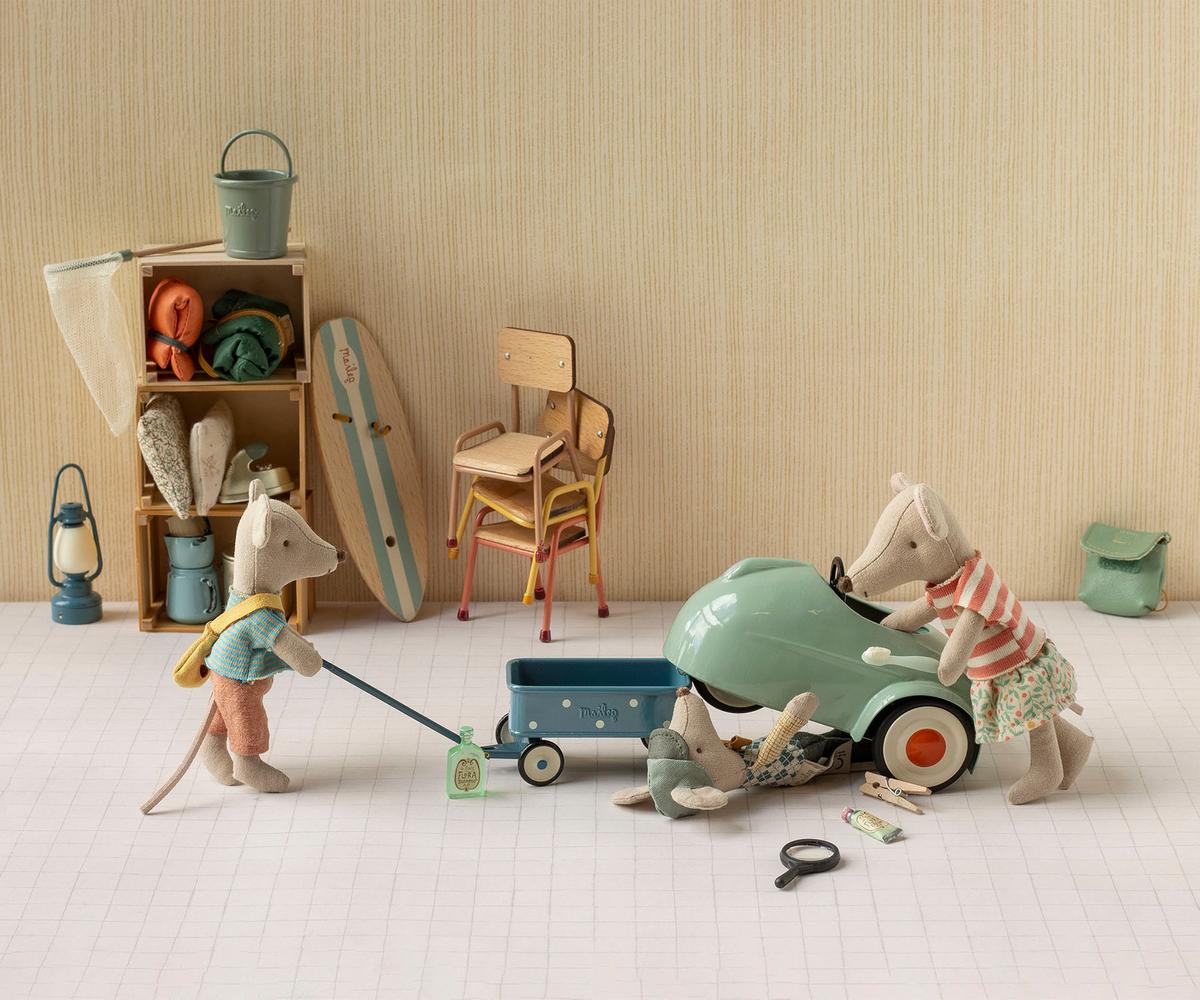 Three Maileg mice in a room with camping gear inspect a Light Mint Car – Mouse; one mouse lies under the car, one looks inside, and one pulls a pull along wagon.