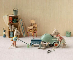 Three Maileg mice in a room with camping gear inspect a Light Mint Car – Mouse; one mouse lies under the car, one looks inside, and one pulls a pull along wagon.