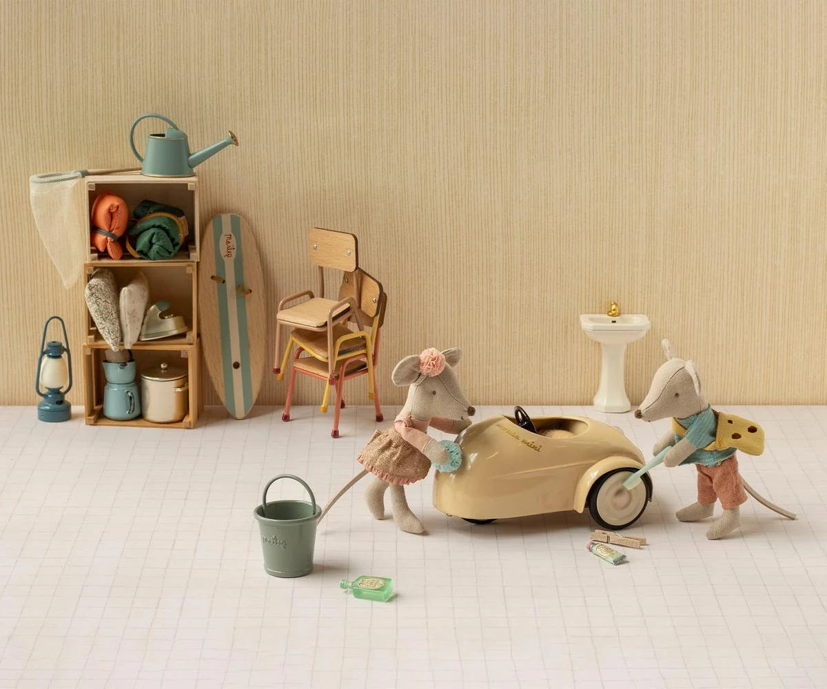 Two Maileg toy mice stand beside a small wheat-colored Car, Mouse in a room furnished with a sink, shelf, chairs, and miniature household items.