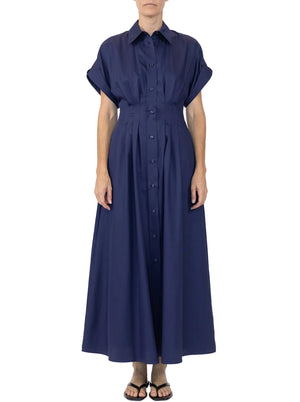 A person stands wearing the Callas Milano Corsica Dress, a navy blue cotton twill, short-sleeve, collared, button-down maxi with a flattering fit and flare silhouette, paired with black sandals.