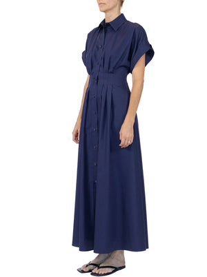 A person wears the Corsica Dress by Callas Milano, a navy blue cotton twill short-sleeve button-down maxi dress with a collar, paired with black flip-flops, standing against a white background.