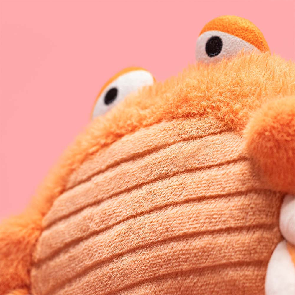Close-up of the Jellycat Pinchsnap Crab, an orange plush toy with big eyes and a ribbed belly, shown against a solid pink background.