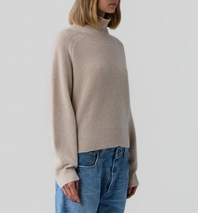 A person wears the Guest in Residence Cropped Ribbed Turtleneck in beige with blue jeans, standing against a plain light gray background.