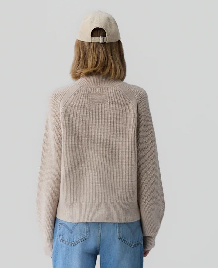 Person facing away from the camera, wearing Guest in Residence’s Cropped Ribbed Turtleneck in beige, blue jeans, and a beige baseball cap, standing against a plain light background.