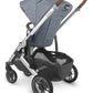 A side view of the UPPAbaby Cruz V2 Stroller by UPPAbaby, featuring a grey fabric toddler seat, a brown adjustable handlebar, and a silver frame. The stroller includes a storage basket underneath and four black wheels.