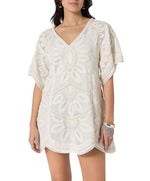 A woman models the Scarlett Poppies Ulay Dress, a short, loose-fitting white dress with embroidery, short sleeves, V-neckline, and artisanal details, styled with a bracelet.