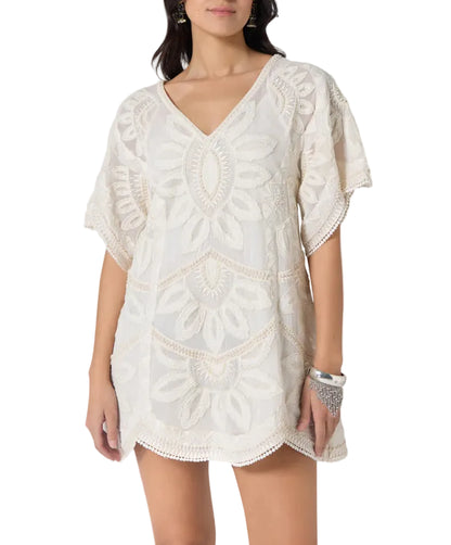 A woman models the Scarlett Poppies Ulay Dress, a short, loose-fitting white dress with embroidery, short sleeves, V-neckline, and artisanal details, styled with a bracelet.