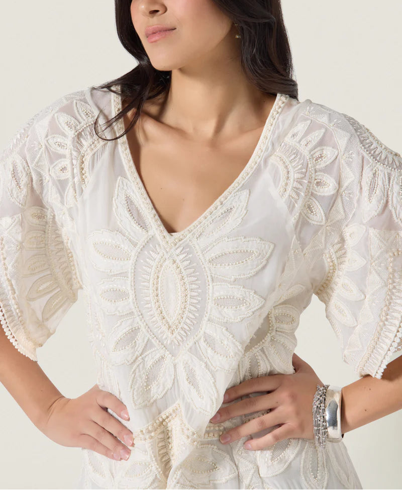 A woman stands with hands on hips, wearing the Ulay Dress by Scarlett Poppies—a white embroidered V-neck dress with floral patterns and artisanal detailing, accessorized with a silver bracelet.