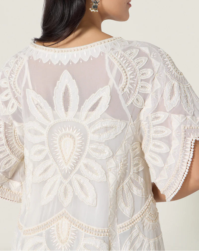 A woman wearing the Scarlett Poppies Ulay Dress, a sheer white blouse featuring embroidered floral patterns, sculptural volume, and scalloped sleeve edges.