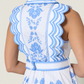 A woman models the Scarlett Poppies Vixeen Dress, a white sleeveless design with blue floral embroidery, scalloped trim, sculptural tailoring, and a belted waist for an elegant silhouette; shown from the back.