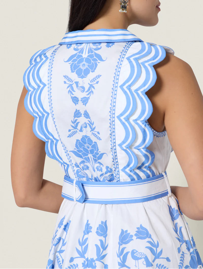 A woman models the Scarlett Poppies Vixeen Dress, a white sleeveless design with blue floral embroidery, scalloped trim, sculptural tailoring, and a belted waist for an elegant silhouette; shown from the back.