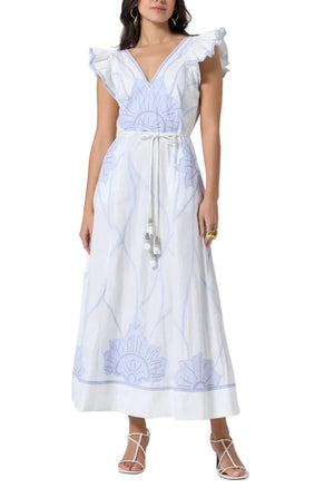 A woman wears the Scarlett Poppies Vittorio Dress—a white sleeveless maxi with a feminine silhouette, ruffle shoulders, blue statement embroidery, tie waist—and pairs it with white strappy sandals.