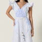 A woman wears the Vittorio Dress by Scarlett Poppies, showcasing a feminine silhouette with light blue statement embroidery, ruffled sleeves, a V-neckline, and a tasseled belt, set against a plain background.