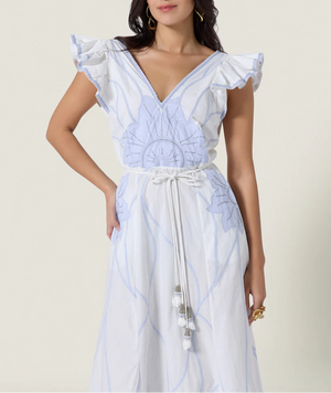 A woman wears the Vittorio Dress by Scarlett Poppies, showcasing a feminine silhouette with light blue statement embroidery, ruffled sleeves, a V-neckline, and a tasseled belt, set against a plain background.