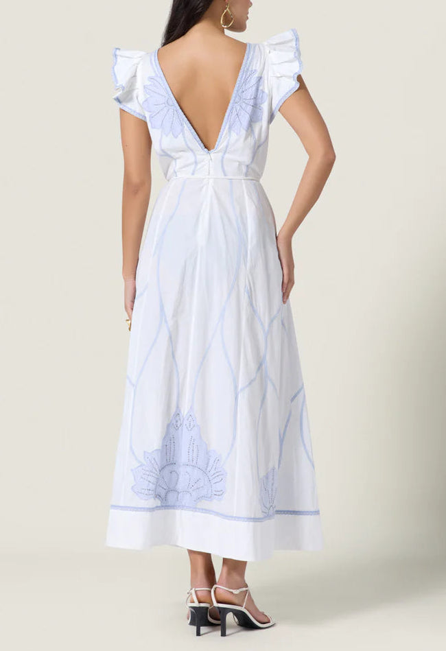 A woman models the Vittorio Dress by Scarlett Poppies, showcasing statement embroidery, ruffled cap sleeves, a deep V-back, and a feminine silhouette. She pairs it with white heeled sandals against a plain backdrop.