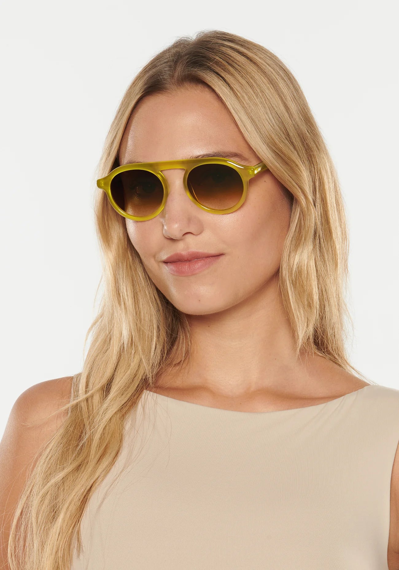A woman with long blonde hair wears Krewe Cameron Sunglasses—yellow acetate frames with round lenses—and a beige sleeveless top, posing against a plain white background.