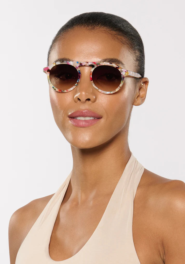 A person models Krewe's Cameron Sunglasses with colorful round lenses and a signature brow bar, paired with a beige halter top, against a plain white background.