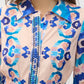 A woman wearing the Figue Cami Dress, featuring a pastel pink base with bold blue and aqua abstract patterns, blue decorative trim along the button placket, and an exaggerated collar.